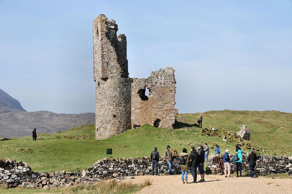 Ardvreck Castle. The image shows the stump of a castle in the centre of the frame, with part of a circular turret on the left and other remains attached to its right. In the foreground is a dry-stone wall and a beach this side of it, with a group of people on the beach and the field beyond the wall. There are other people by the castle. The sky is blue.