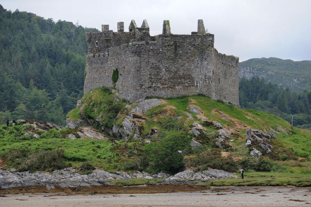Castle Tioram. The image shows the irregularly shaped curtain wall of a stone castle standing on top of a tidal island with a shoreline in the foreground. A person is standing in the bottom right of the frame. The wall heads of interior buildings can be seen projecting above the top of the curtain wall. In the background is a hillside covered in conifer trees. The light is subdued.