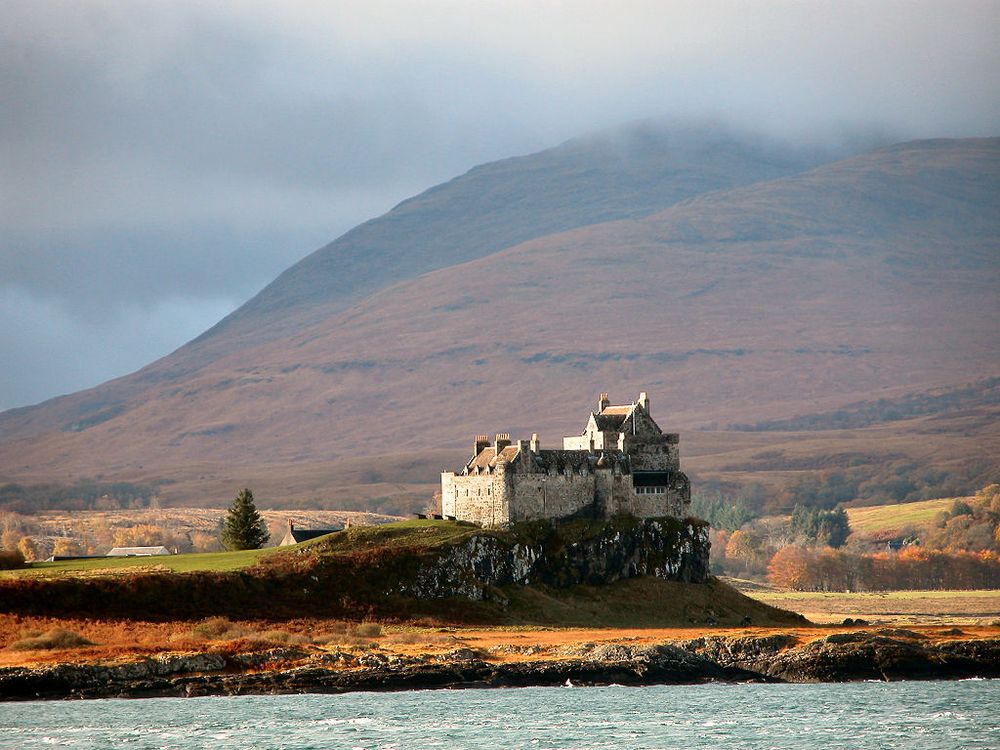 Duart Castle. The image shows the castle standing on a rocky outcrop seen from the sea, with the shore in the foreground. The vegetation is very brown and, in the background, mountains rise into the clouds. The scene is in low sunlight coming from the left.