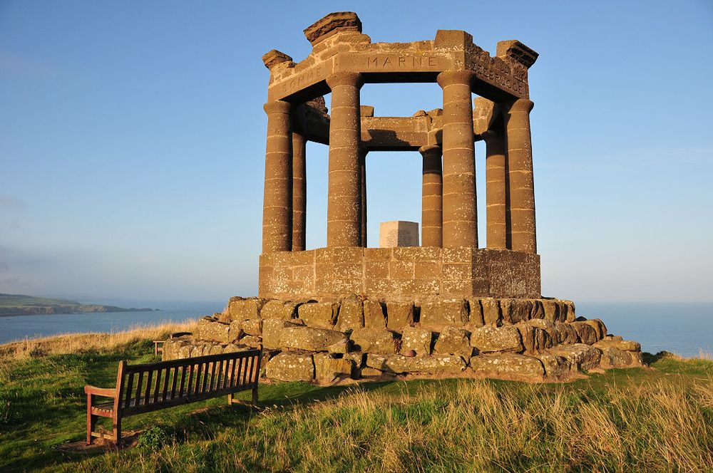 Stonehaven's war memorial. The image shows an open octagonal stone structure with its top supported by round columns. It is set on a three-tiered rough stone base. There is a bench to the left of the memorial and the sea can be seen at a much lower level in the background. The scene is in strong sunlight.