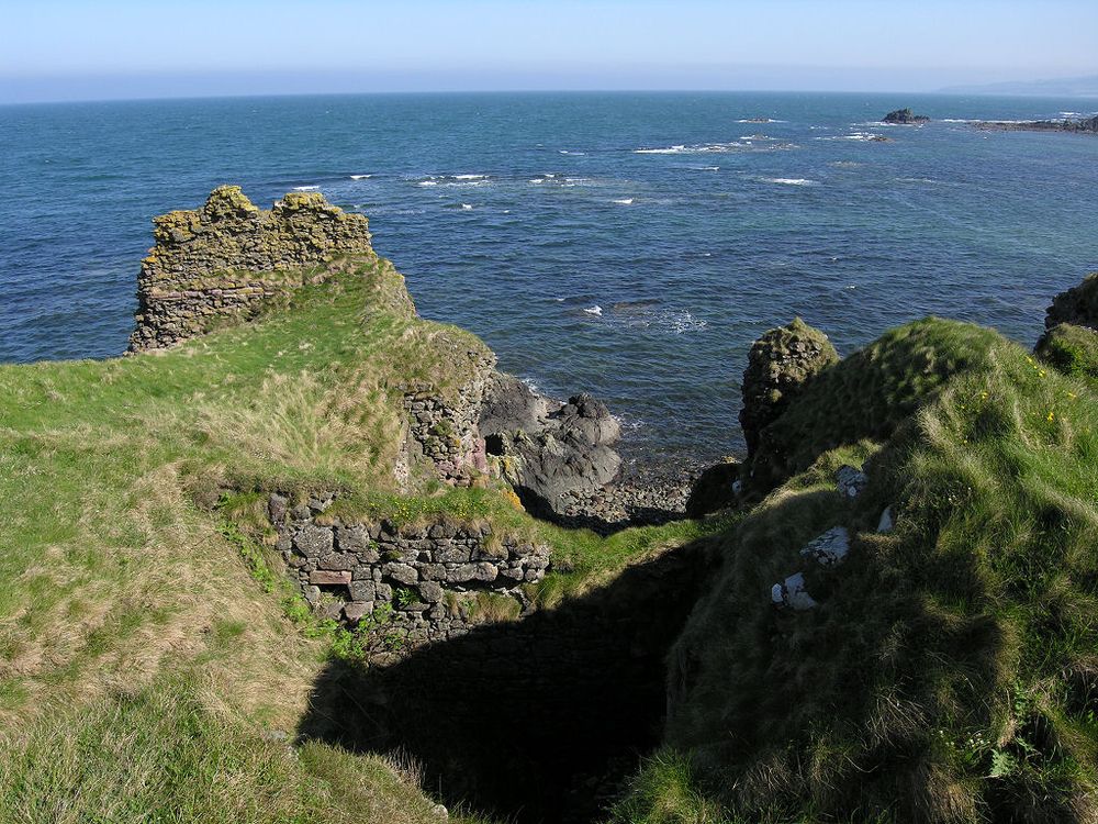 The fragmentary remains of Turnberry Castle. The image shows a view down over rough grass to a shore with the blue sea beyond it. A narrow valley in the centre of the frame has traces of stonework either side of it and there’s part of a stone wall still standing on a short promontory in the upper left of the frame. The scene is in sunlight.