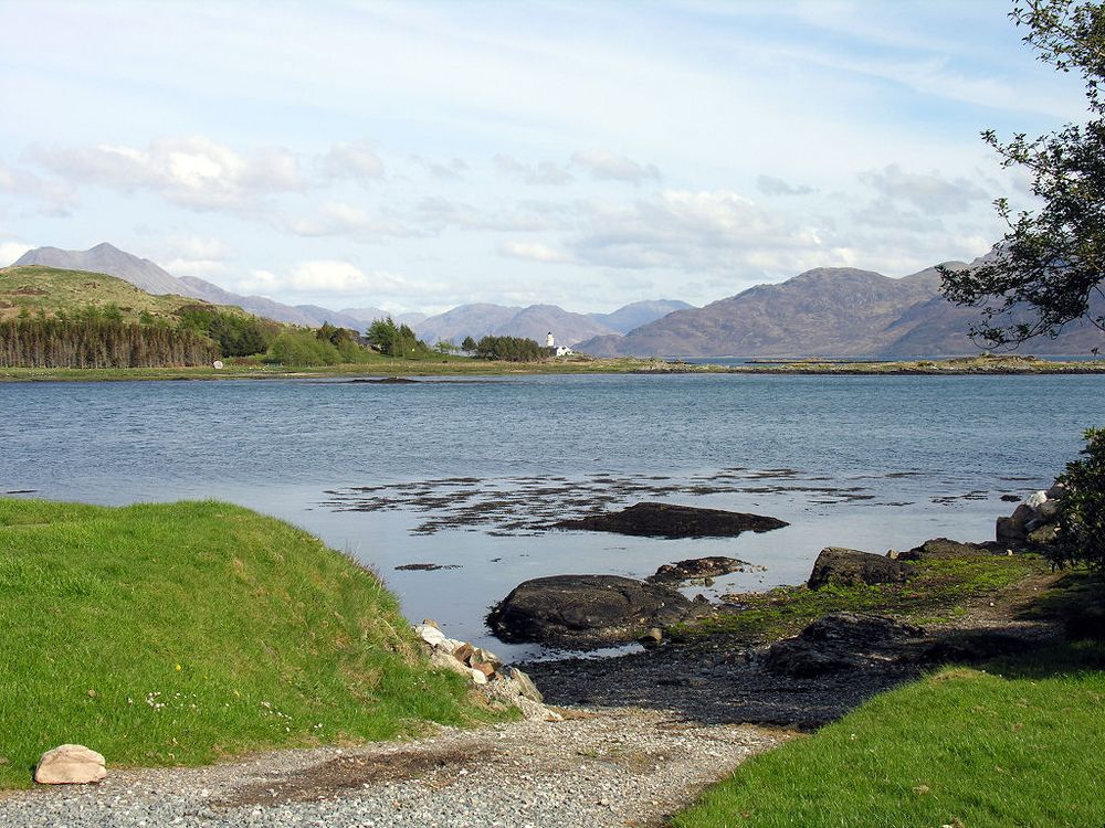 The view east towards the island of Ornsay and its lighthouse. The image shows a gravel slipway leading down into a still sea, with grassy banks on both sides and part of a tree on the right. In the middle distance is part of an island with trees and, just visible, a lighthouse. In the background beyond more sea is a mountainous landscape. The scene is in sunlight.