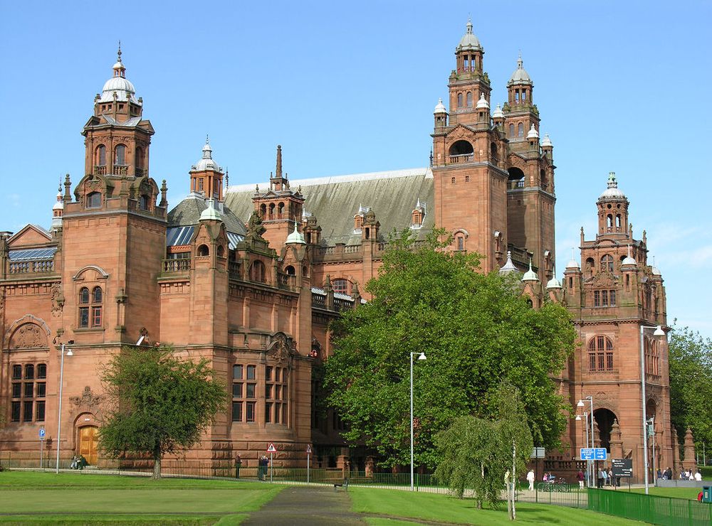 Kelvingrove Art Gallery and Museum in Glasgow. The image shows a very large red stone building topped off with pinnacles and rounded towers. It is facing diagonally to the right. In the foreground a path leads across grass towards the building. The sky is blue.