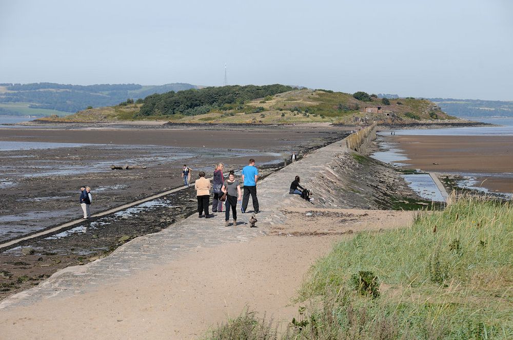 Cramond Island. The image shows the causeway leading to Cramond Island in the Firth of Forth near Edinburgh at low tide, with a group of people starting to cross and the island in the distance. The concrete causeway extends from the bottom left to the upper right of the frame and the island stretches across the picture, with mud this side of it. The scene is in sunlight.