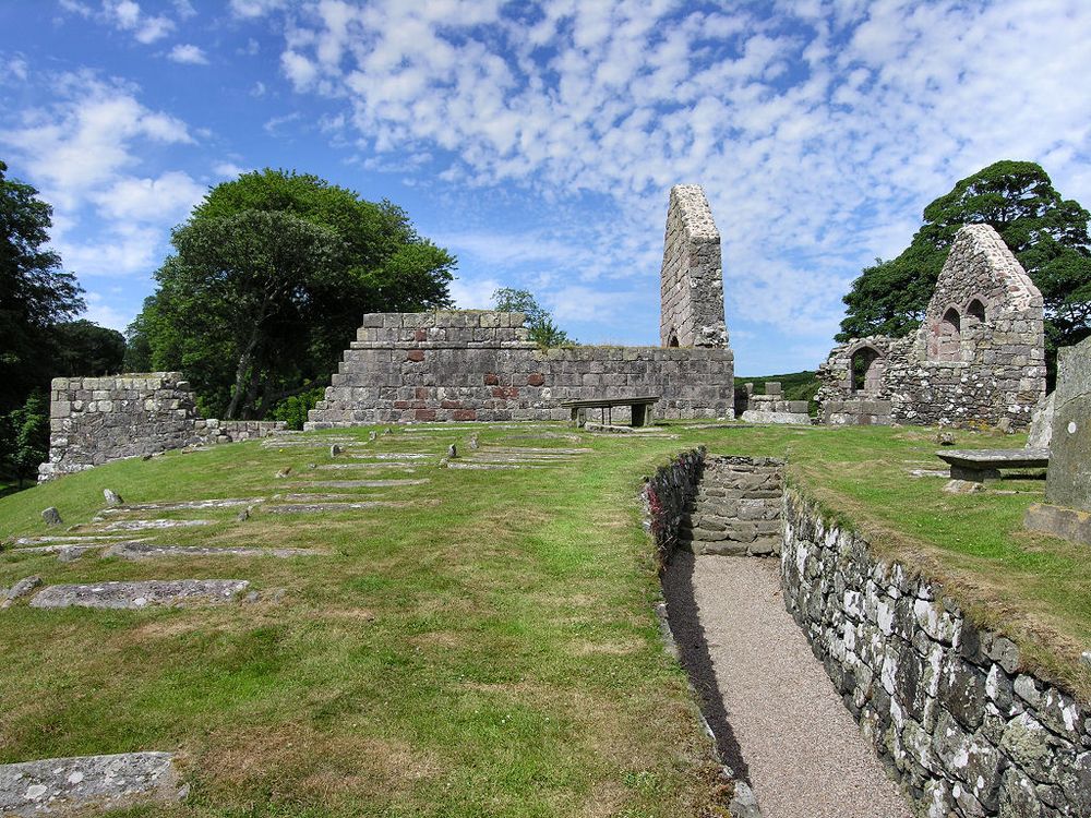 The ruins of St Blane's Church near the southern end of the Isle of Bute. The image shows a path leading slightly uphill in the bottom right of the frame, cut into the ground and flanked by stone walls and then climbing steps. We are looking up to the long side of the ruined church on the skyline, with two standing gables. There are gravestones in the lower left of the frame lying flat on the ground, The scene is in sunlight.