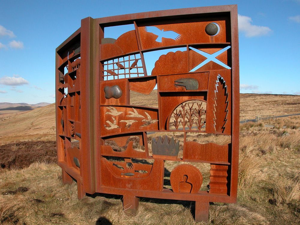 The Hugh MacDiarmid memorial. The image shows a brown bracken hillside. Dominating the picture is a large rust-coloured memorial shaped like an open book facing off to the right. It is decorated with cutouts and patterns. The sky is largely blue.
