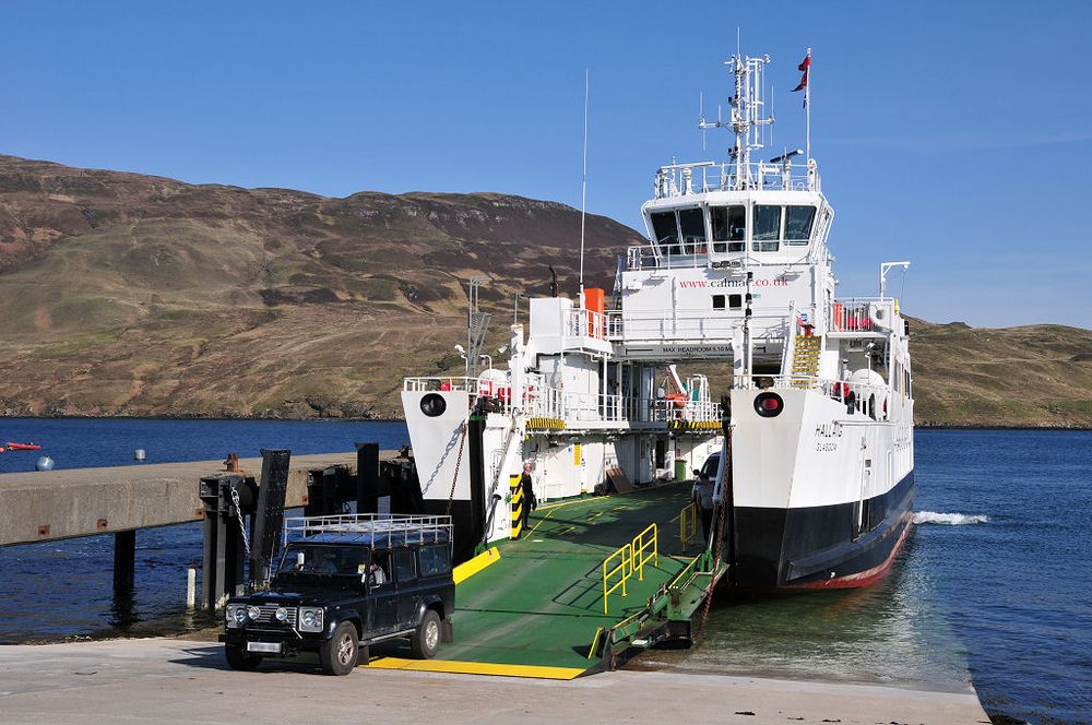 The Isle of Raasay ferry, MV Hallaig. The image shows a broad concrete slipway at the foot of the frame with a white and black ferry berthed end on to the camera with its bow door down and a black Land Rover driving off. There is sea in the background and then a hillside covered in brown bracken. The sky is blue.