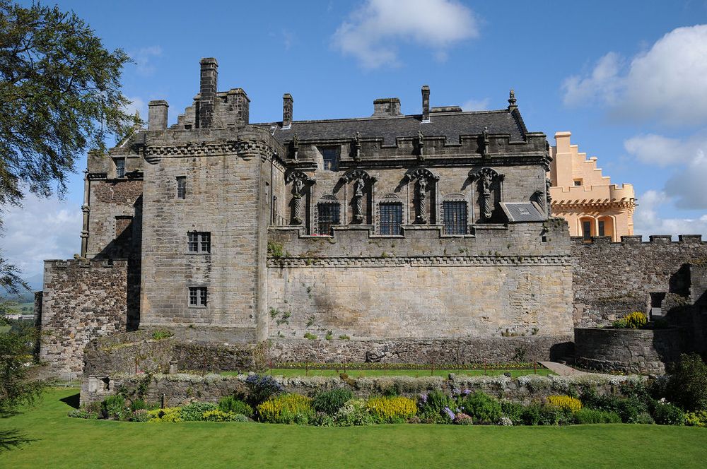 The palace at Stirling Castle, seen across its garden. The large grey stone palace stretches across much of the centre of the frame, with the bright harling of the great hall visible to its right. In the foreground are formal gardens and a lawn while part of a tree is visible on the left. The scene is in sunlight.