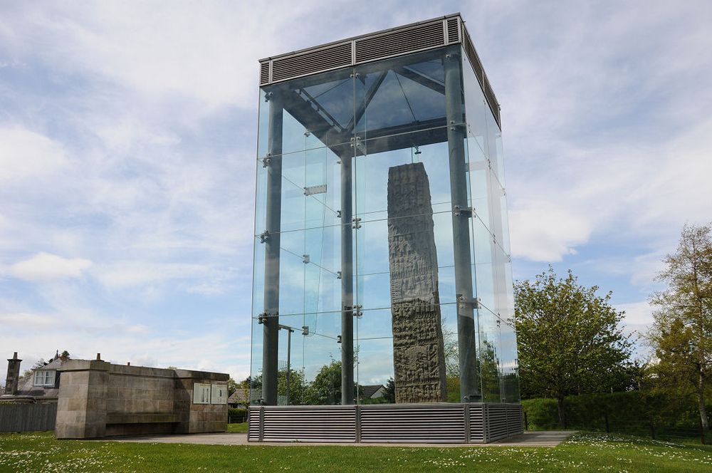 Sueno's Stone. The image shows a view from slightly below of a tall transparent box supported by metal corner pillars and frame. Within the box is a tall carved stone with row upon row of carved figures in a very complex arrangement. There’s a low wall to the left with a seat and houses in the background. The scene is in sunlight.