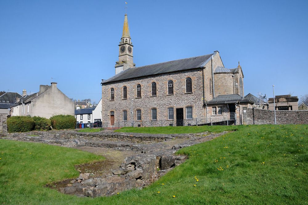 Lesmahagow Priory in South Lanarkshire. The image shows a stone church standing in the middle distance with a narrow spire at its left-hand end. There are houses and other buildings in the background. In the foreground is a grassy area with the remains of stone foundations visible slightly above and below the ground level. The sky is blue.
