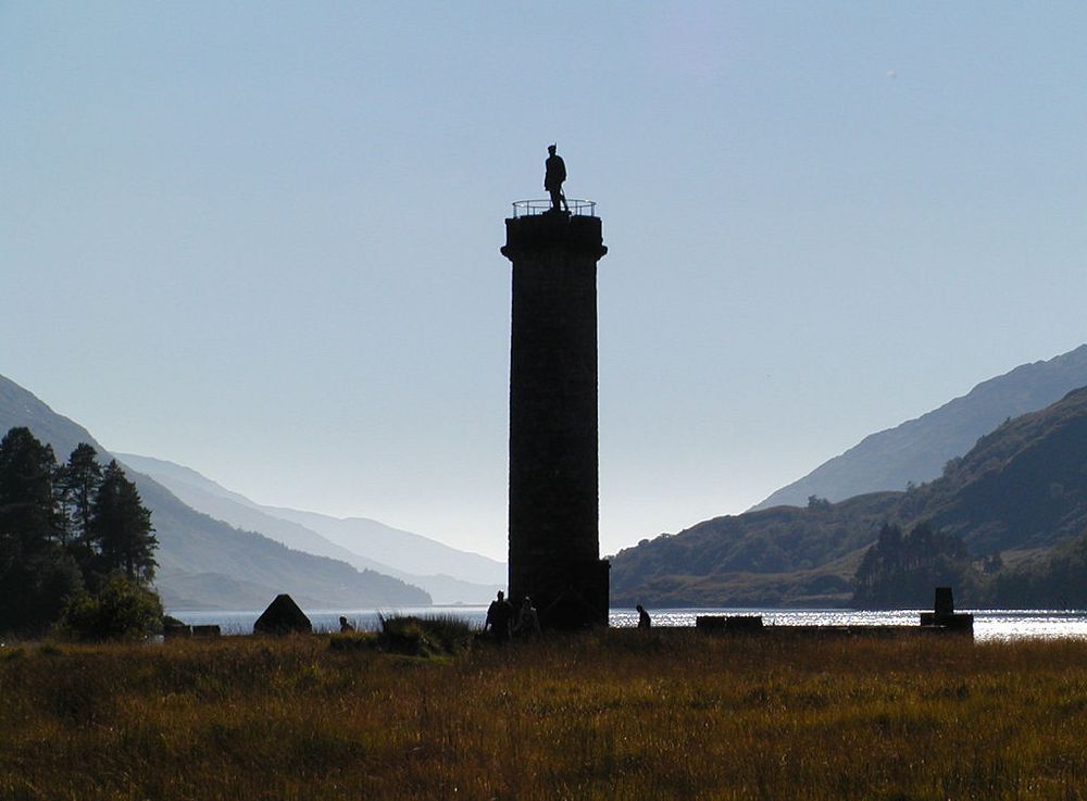 The Glenfinnan Monument. The image shows a view over rough ground to the low walls surrounding a tall column with the figure of a Highlander on the top of it. The monument is silhouetted by light from behind it. In the background is a large loch extending away from us, with hills rising on both sides. The sky is blue.