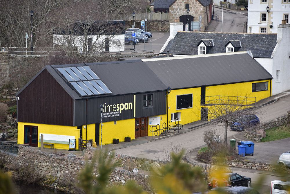 Timespan Museum and Arts Centre. The image shows a view down to an oblong building that extends from bottom left of the frame into the middle right. There is a road climbing a hill this side of it and other buildings and a car park in the background seen above the building, which is mainly yellow with dark brown facings at its left-hand end. The light is grey.