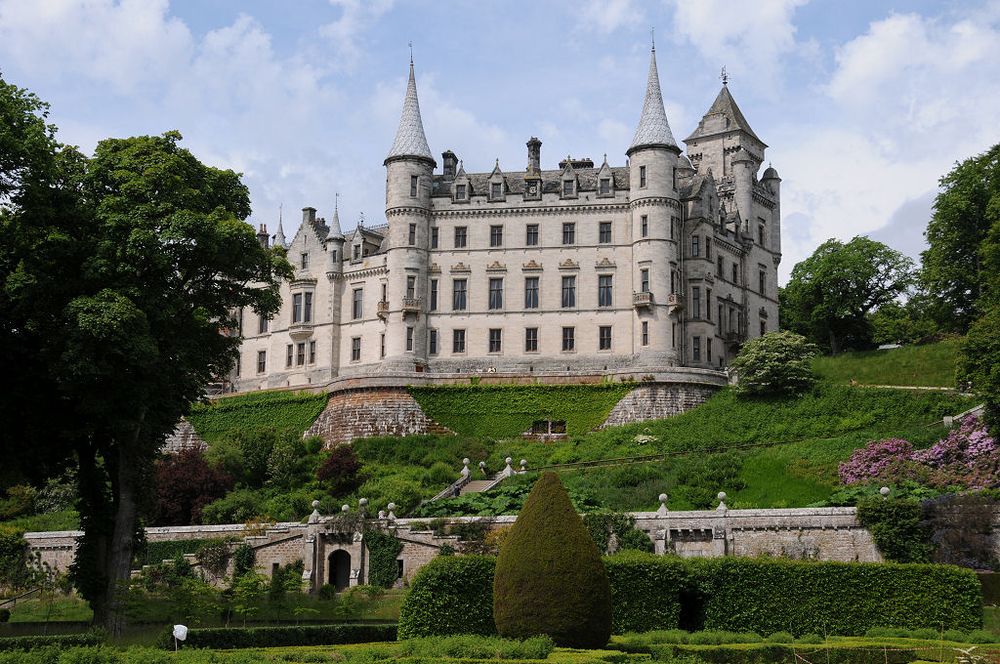 Dunrobin Castle near Golspie in Sutherland. The image shows formal gardens in the foreground which lead back to a steep slope on top of which is a fairy tale style castle in light grey stone. It is up to four storeys in height and topped off with turrets and ornaments. There’s a tree in the left foreground, The light is subdued.