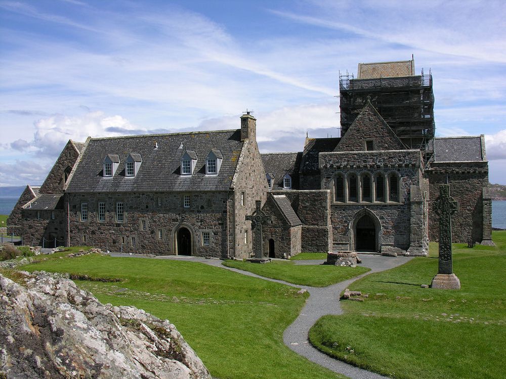 Iona Abbey. The image shows an abbey with sea in the background. The abbey church is seen end-on on the right and other buildings are to its left. There is mown grass with paths in the foreground and an outcrop of rock on the lower left of the frame. The scene is in sunlight. 