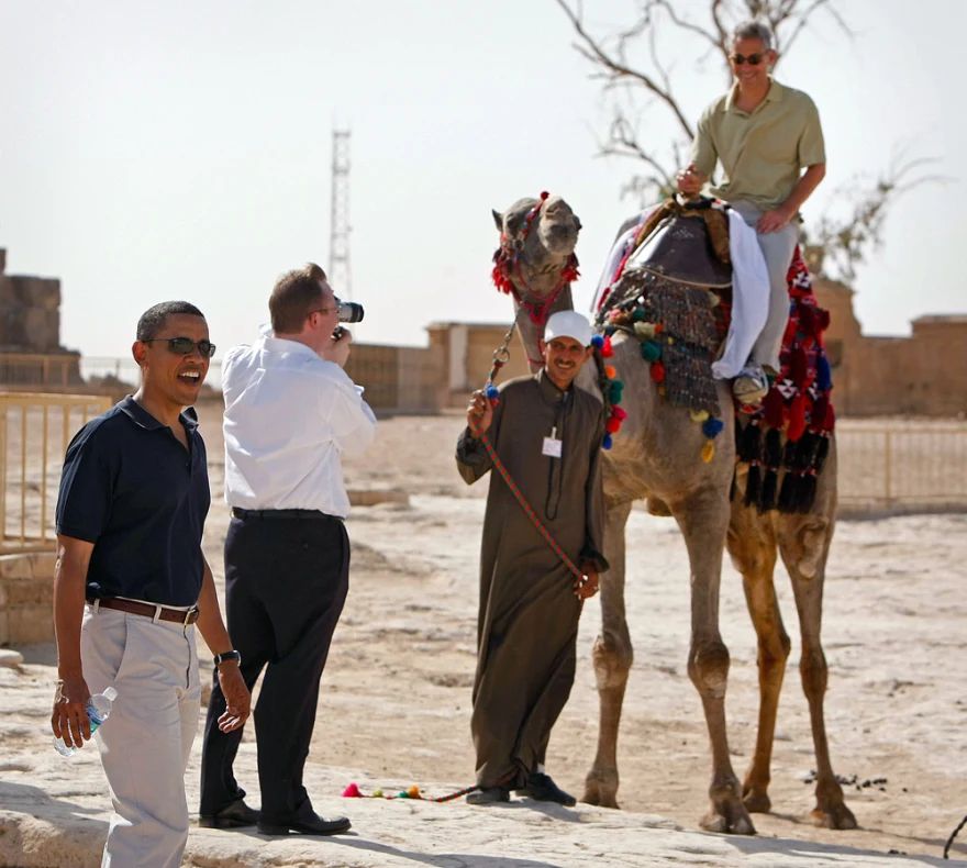 (June 2009, Egypt) Barak Obama, a photographer, and a camel handler stand while Rahm Emmanuel sits atop a camel. Rahm thinks the camel must be a male; when he rides it, people laugh and exclaim 