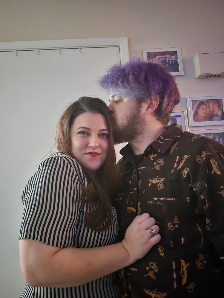 Katie, a woman with auburn hair in a striped dress, embraces Steve, a man with purple hair in a patterned shirt. Steve is kissing Katie's head. 