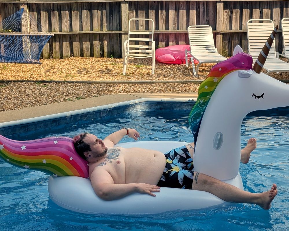 A chubby white person lounges on a unicorn shaped inflatable pool raft in a pool. They are only wearing grey swim trunks with blue and yellow Palm trees. 