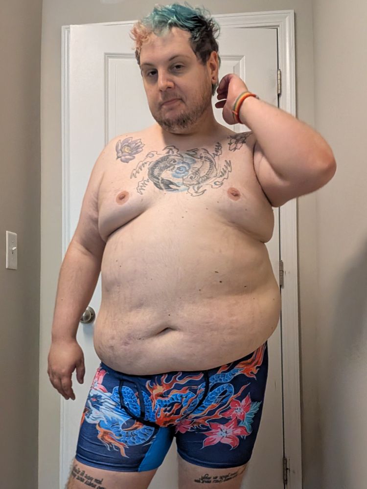 A chubby white person standing in front of a door. They are wearing only underwear. They have scruffy facial hair, tattoos on their chest and thighs. The underwear are a deep blue with a dragon fire and flowers. 