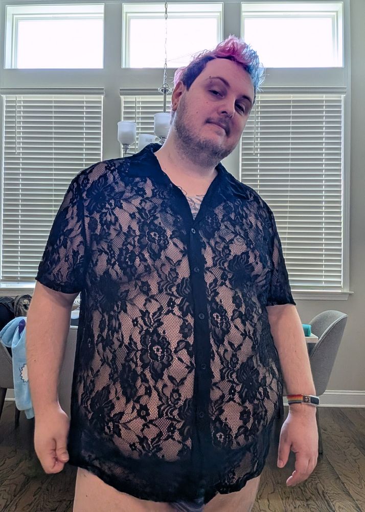 A chubby white person in a black colored knit button down shirt. The knit has lots of gaps allowing the skin under the shirt to be visible. The not pattern looks like flowers.  They also have blue stripes briefs on.