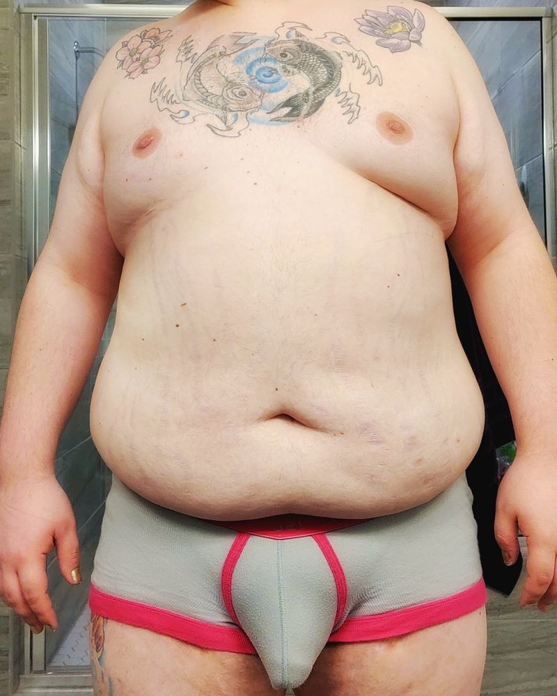 A photo of a chubby white person wearing only underwear. The image starts at their shoulders and ends just above the knees. The underwear is grey with pink piping. Their bulge is very prominent with "VPL"