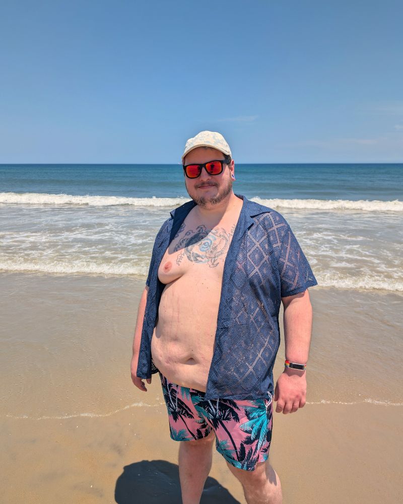 A fat white person on the beach. They have a sheer navy colored shirt on that's open to the wind exposing their large belly. They're wearing  Pink and blue swim trunks with palm tree silhouettes. 