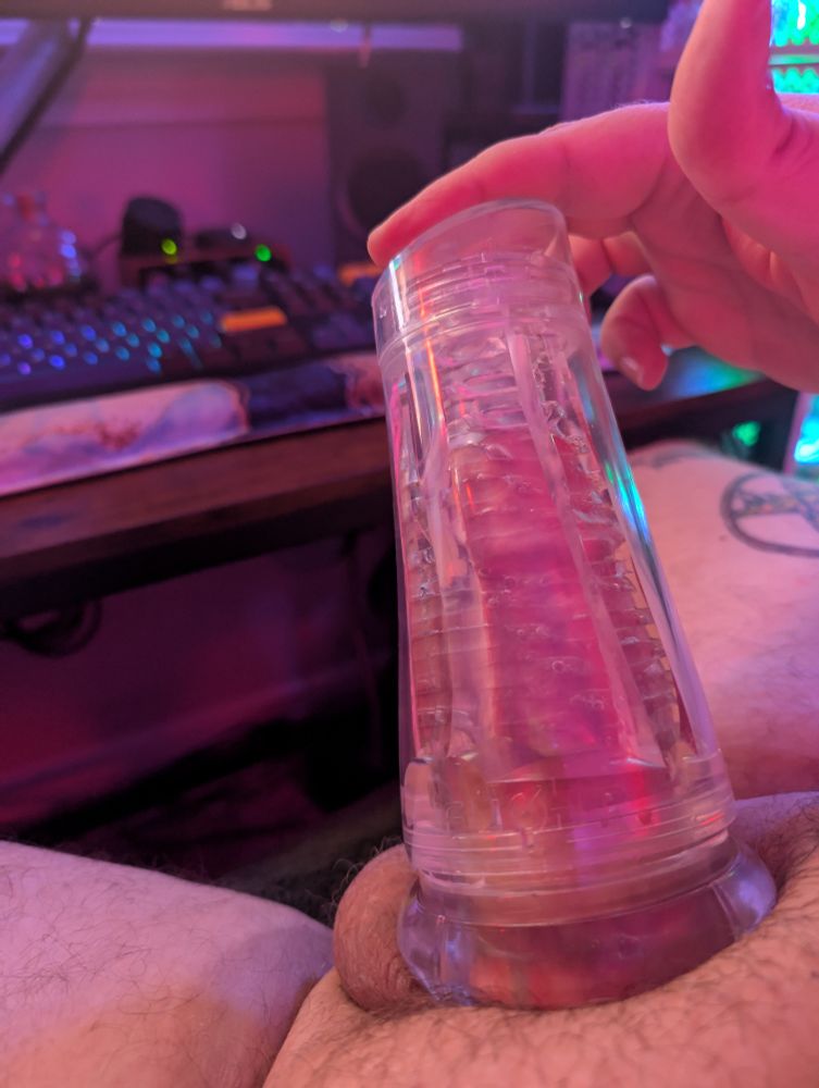 A photo of a white person's erect penis fully inserted into a clear masturbation device. In the background is a desk and keyboard. 