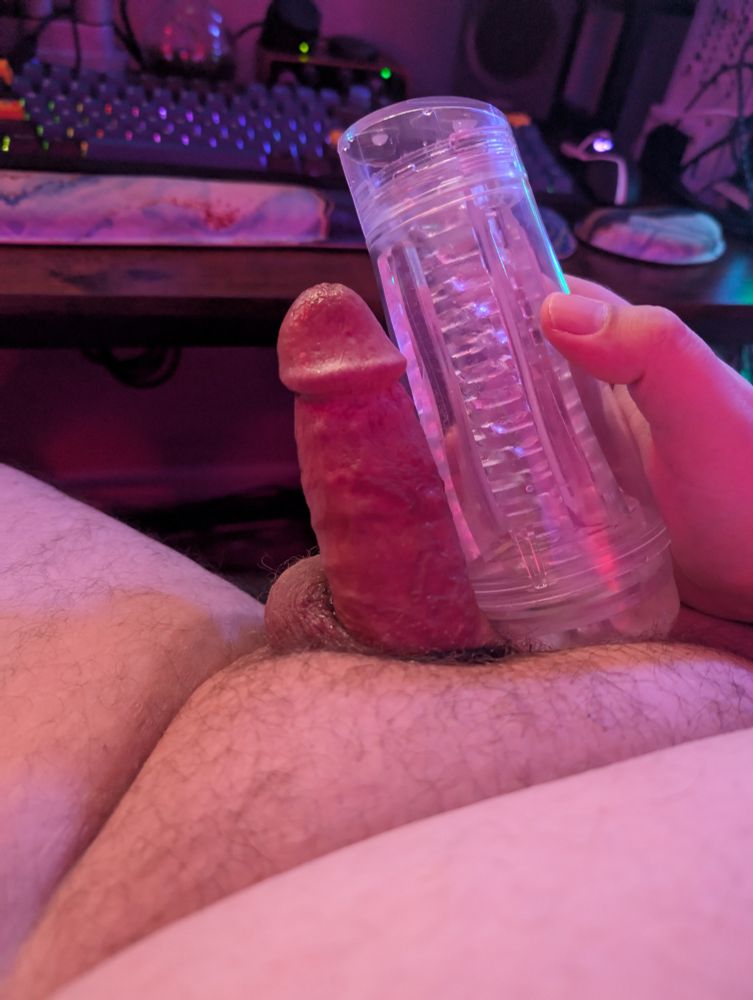 A photo of a white person's erect penis next to a clear masturbation device. In the background is a desk and keyboard. 
