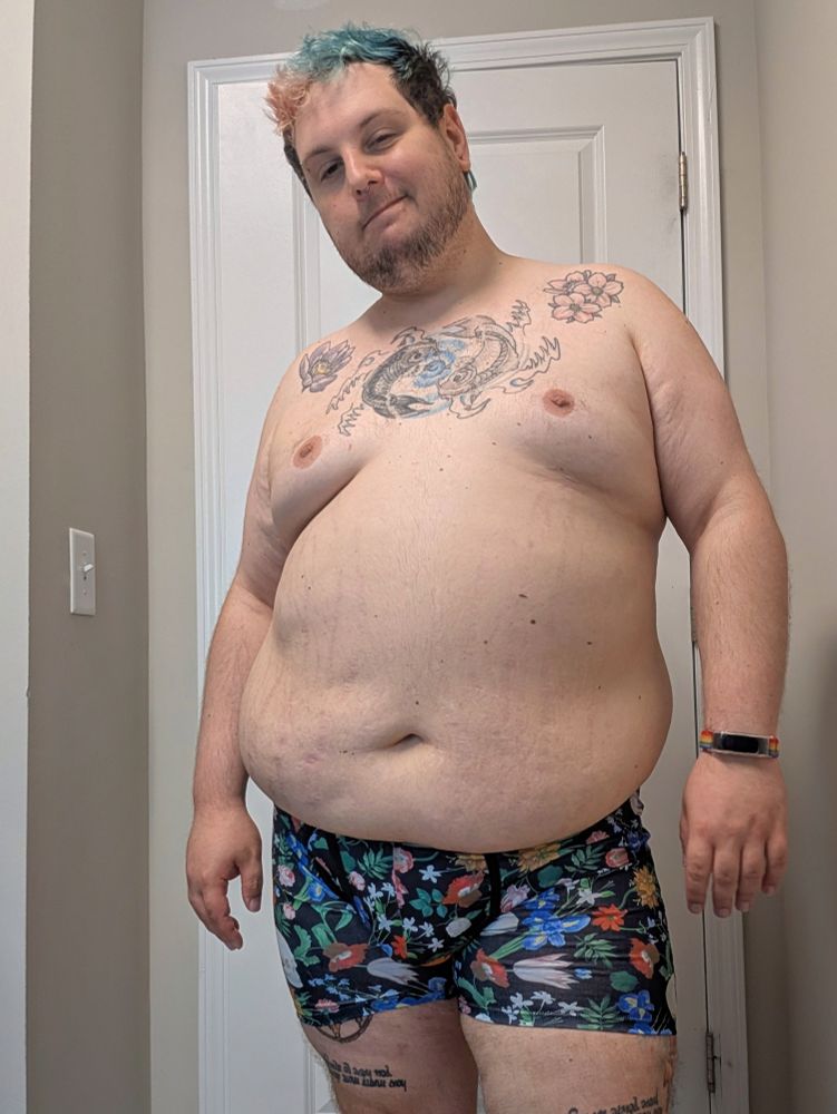 A chubby white person standing in front of a door. They are wearing only underwear. They have scruffy facial hair, tattoos on their chest and thighs. The underwear are black with colorful flowers