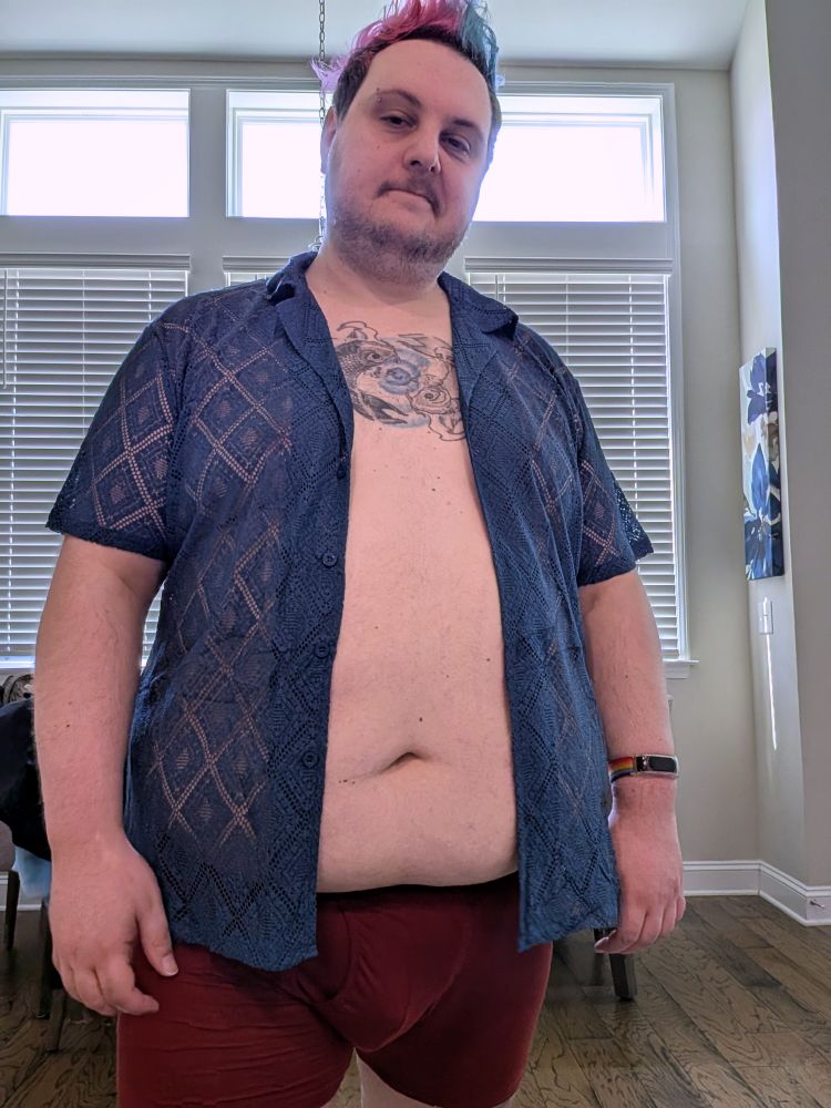 A chubby white person in a navy colored knit button down shirt. The knit has lots of gaps allowing the skin under the shirt to be visible. The shirt is unbuttoned showing off more of their chest and belly. They also have dark red underwear on. 