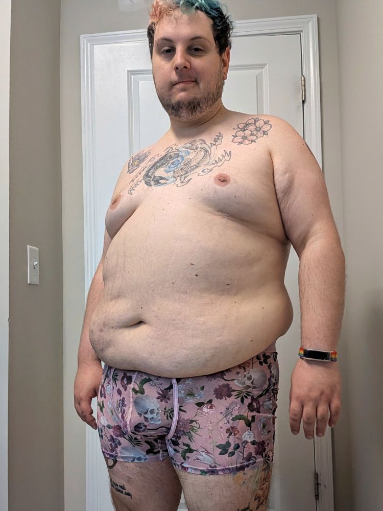 A chubby white person standing in front of a door. They are wearing only underwear. They have scruffy facial hair, tattoos on their chest and thighs. The underwear are pink mesh with skulls and flower designs. 
