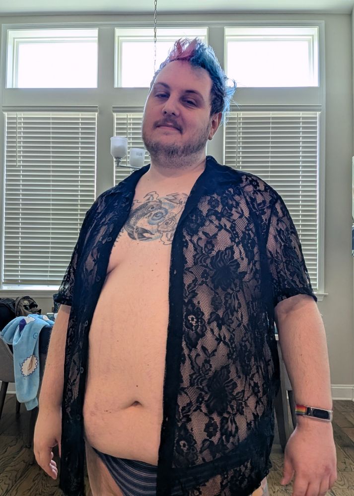 A chubby white person in a black colored knit button down shirt. The knit has lots of gaps allowing the skin under the shirt to be visible.  The shirt is open exposing their belly. They also have blue striped briefs on.