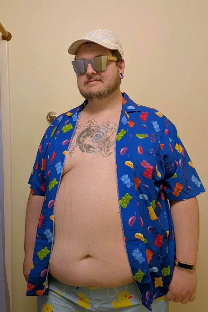 A chubby white person with a beard stands in front of a pale yellow wall. They have on a canvas hat, silvery mirror shades, a royal blue shirt with the pattern of jelly beans and gummy bears on it and a pair of swim trunks with ducks and bubbles. The shirt is open exposing their chest tattoo of two koi fish and their large belly. 
