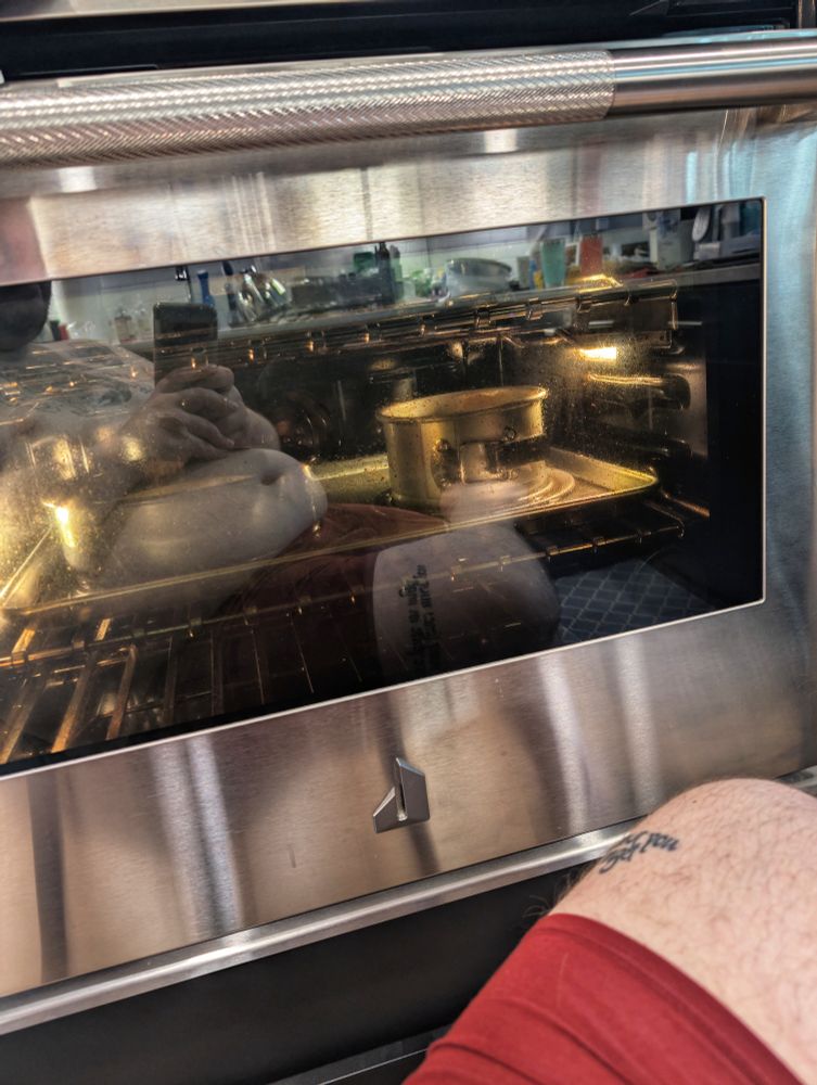 A stainless steel oven with the light on inside. There are two cheesecakes inside the oven but one is obscured by the reflection of a shirtless white person's belly. 