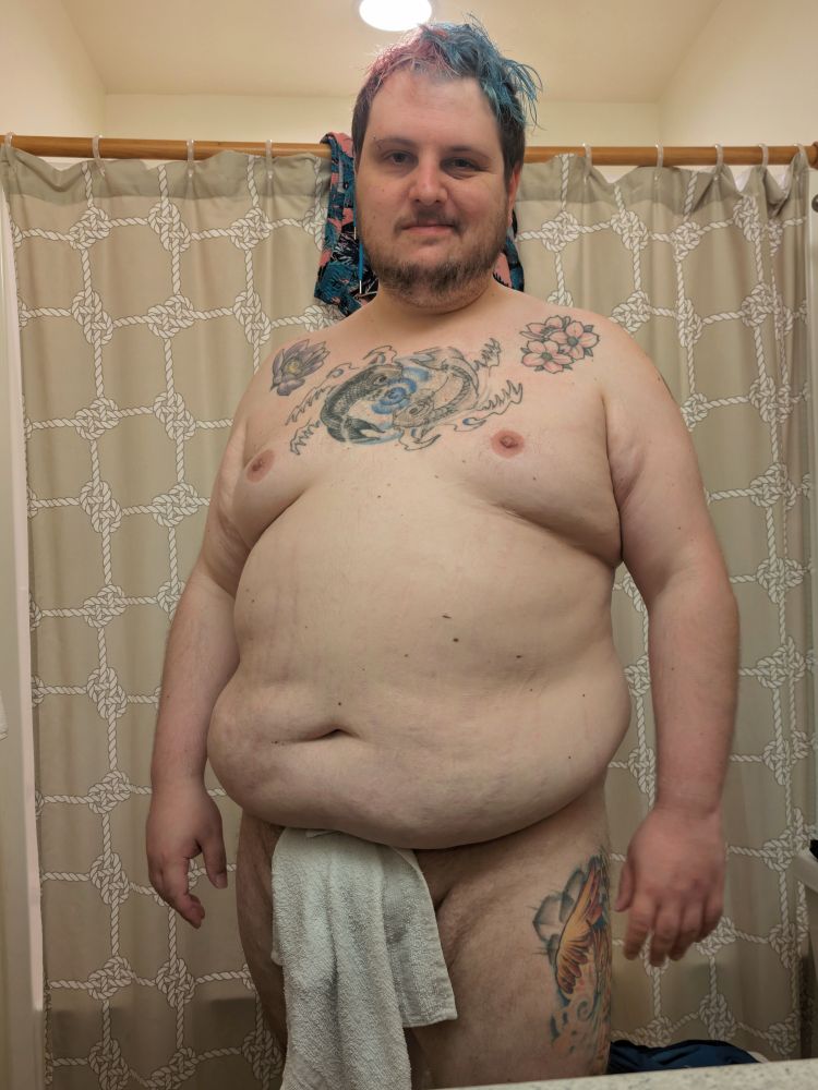A photo of a chubby white person who stands nude in front of the camera. A white towel is held in place by their belly and covering and their genitals. Their hair is pink and blue and messy. Behind them is a beige sailor's knot themed shower curtain 