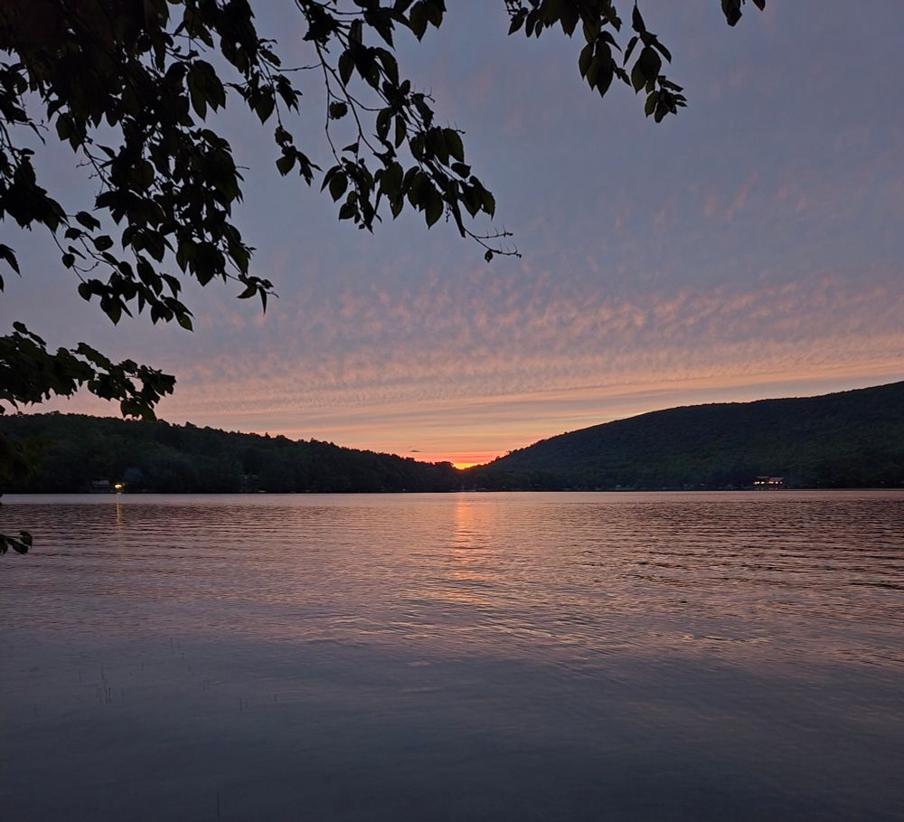 Sunset over a lake with the sun dipping into a notch between 2 hills and a beautiful dappled mackerel sky. Some leaves from a birch tree are in the foreground 