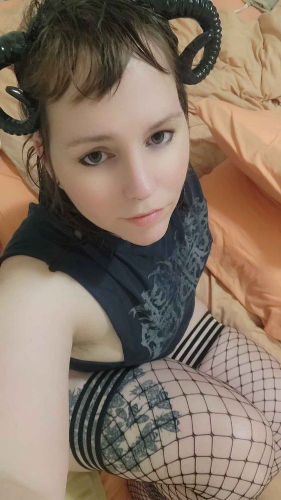 a person with ram-style horns, wearing a dark sleeveless top and fishnet tights with striped thigh-highs, sitting on a bed with orange bedding, looking up at the camera with a calm expression.