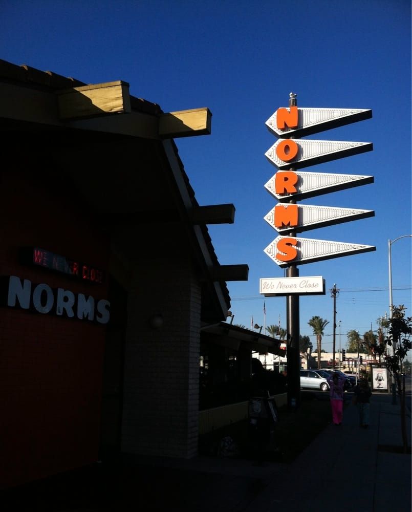 This image shows the iconic retro signage for "Norms," a well-known diner chain, with its arrow-like design and bold, orange letters. It also includes the tagline "We Never Close," reinforcing the 24/7 availability of the diner.