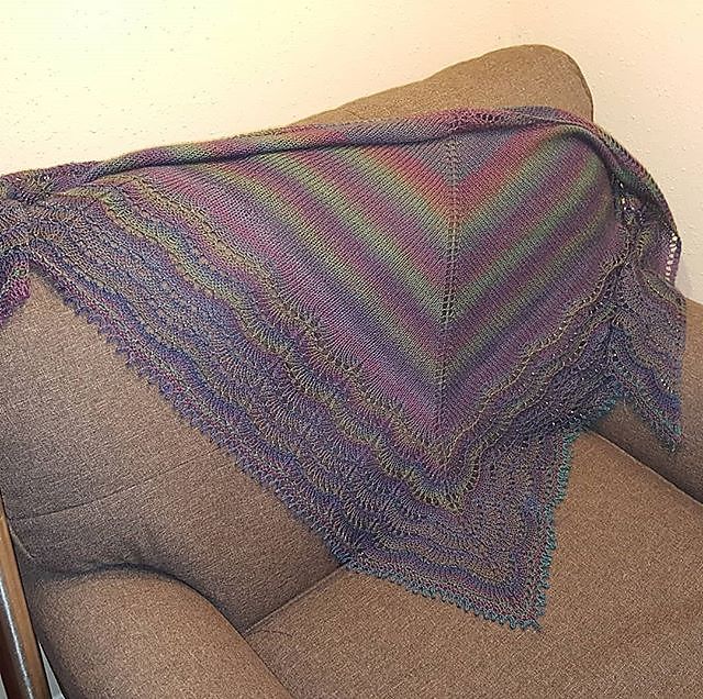A knitted Holden Shawl, with a main body in stockinette and a feather and fan border. The yarn shifts through shades of purple, blue, and green. 