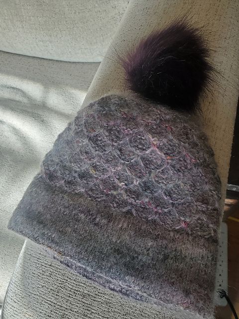 A textured purple knit hat with a pompom on top. The body of the hat is blown out by grey-leaning suri yarn held double with a purple ombre, worked in a slip stitch texture. 