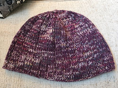 A scrappy hat knitted with two yarns held doubled and marled. One yarn is burgundy, and the other is an ombre with shades of pink, Grey, and a bit of yellow. 