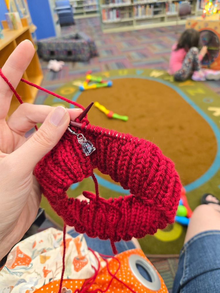 A red ribbed hat brim with a stitch marker visibly marking the beginning of the row. Blurred in the background is a child playing on the floor, back to the camera. 