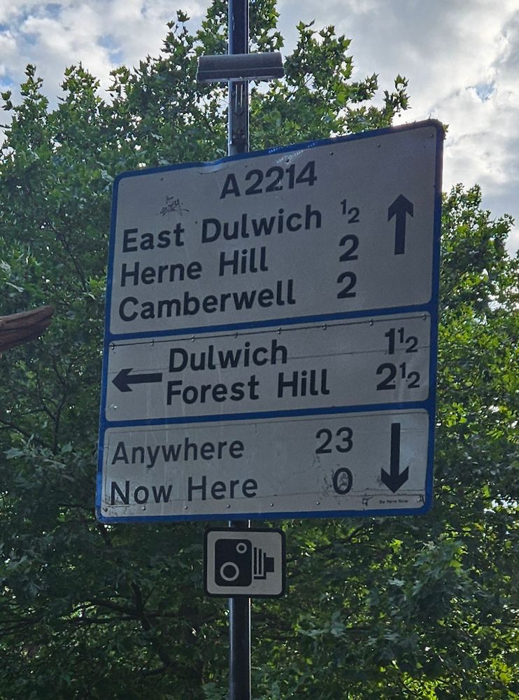 A roadsign that has been tampered with so, alongside directions to East Dulwich (half a mile straight ahead) and Forest Hill (2 1/2 miles to the left), there is an arrow sending you back the way you came to "Anywhere" (23 miles) and "Now Here" (0 miles)