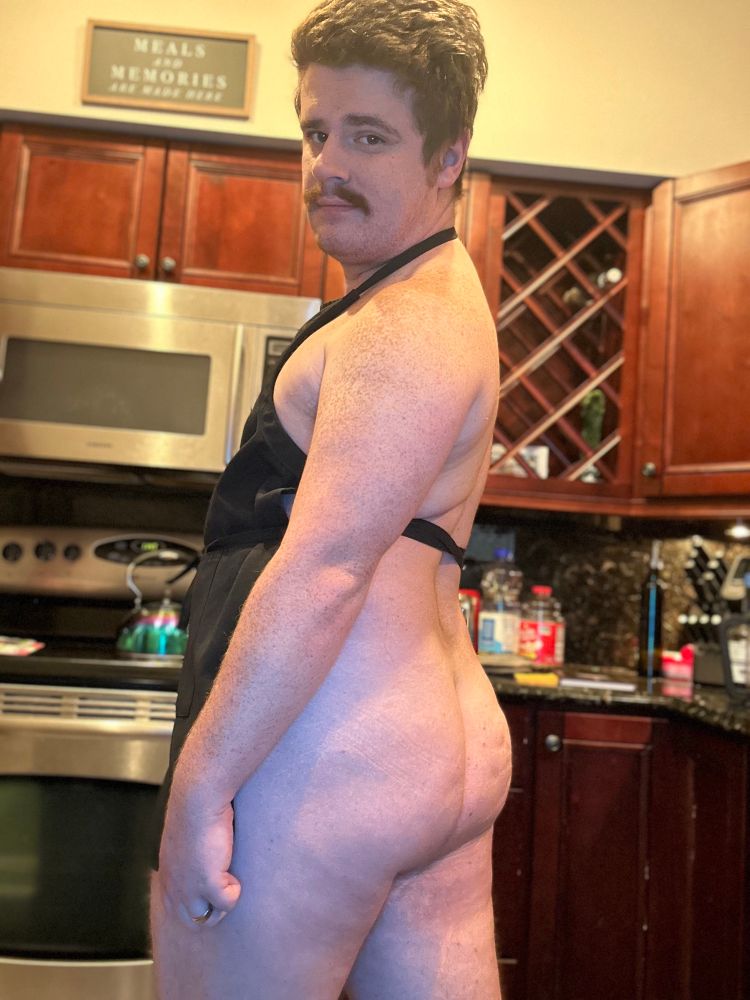 Photo of me standing naked in the kitchen with nothing on but an apron. I’m turned to the camera to show off my round ass