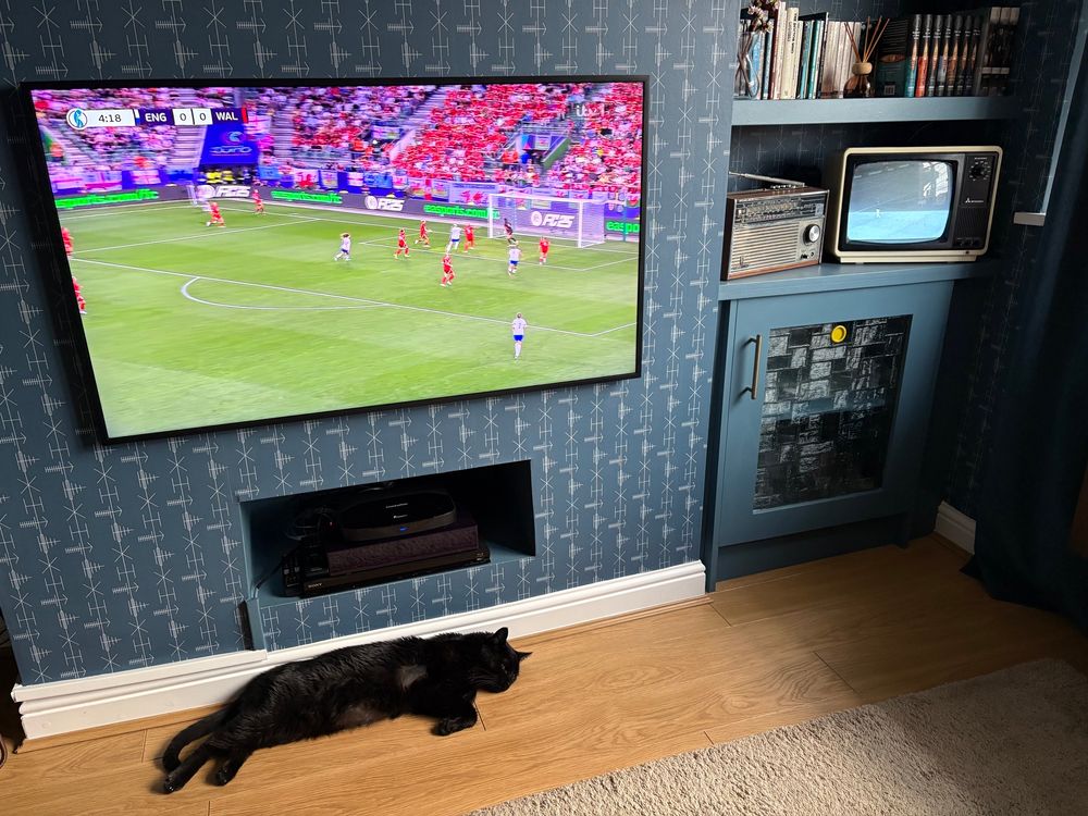 A modern widescreen tv and a 1980s B&W portable both show football. A cat lies asleep on the floor.