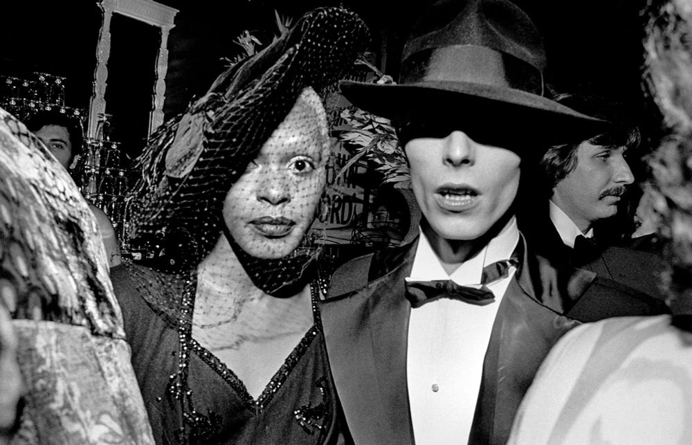 A black-and-white archival photograph by Bill Cunningham capturing two elegantly dressed individuals at a glamorous event. The person on the left wears a veiled hat and a sequined dress, exuding a mysterious allure. The person on the right, dressed in a tuxedo with a bow tie and a tilted fedora, embodies sharp sophistication. Both figures are surrounded by a lively, crowded atmosphere, showcasing the vibrant and eclectic fashion scene of the era.