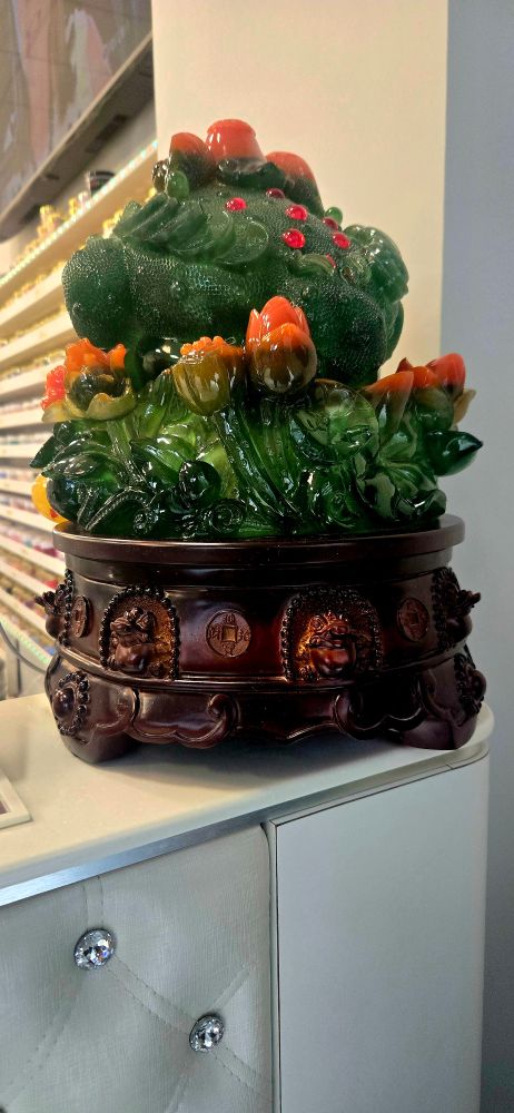 Ornate jade-like sculpture on a wooden base with intricate carvings, featuring green foliage and orange accents. Placed on a cabinet, evoking elegance.