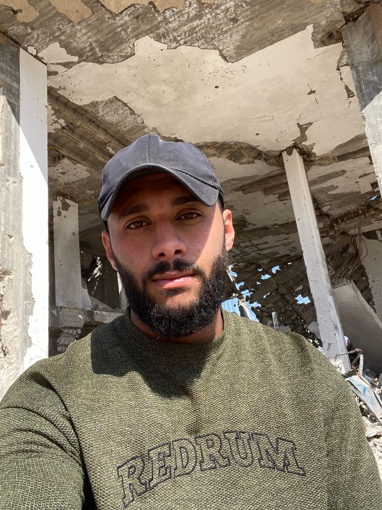 A young man from Gaza stands in front of a completely destroyed building, his face weary but strong. His eyes carry the weight of hunger, fear, and exhaustion—yet he remains standing amid the ruins. Behind him, shattered concrete and twisted steel speak of war, destruction, and loss.
