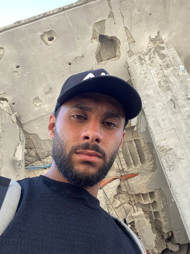 A young man from Gaza stands in front of a bombed concrete wall. His eyes reflect exhaustion, grief, and quiet strength. The ruins behind him speak louder than words. 