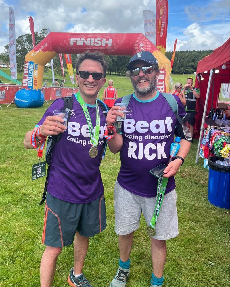 Photo of two white men, smiling at the camera and holding clear cups of water in their hands. They are standing on a grassy field, with an inflatable arch behind them that reads 'Finish'. There are also various flags and tents behind them too. The two men wear dark purple Beat-branded t-shirts. The man on the left has brown hair, wears black-tinted sunglasses and has a medal around his neck with a neon-green ribbon. The man on the right wears reflective sunglasses and has a baseball cap on his head, as well as a grey beard.