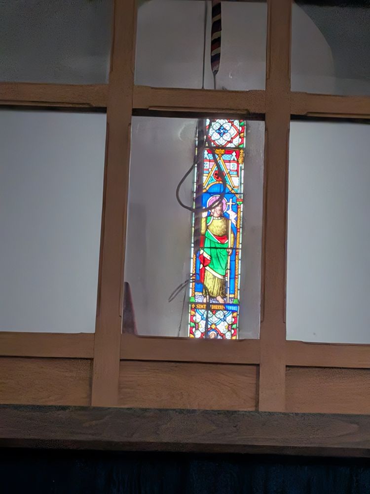View into a bell tower through a window, with the rope looped up like a noose. Behind it a stained glass of S John the Baptist  